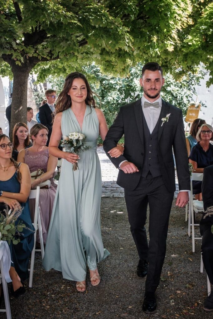 Hochzeitskrone Ein Mann und eine Frau gehen Arm in Arm einen Gang im Freien entlang, der von sitzenden Gästen gesäumt ist, und fangen bezaubernde Momente ein. Die Frau in ihrem hellgrünen Kleid hält einen Blumenstrauß, während der Mann einen dunklen Anzug mit Fliege trägt, perfekt für natürliche Hochzeitsfotos in Niederösterreich.