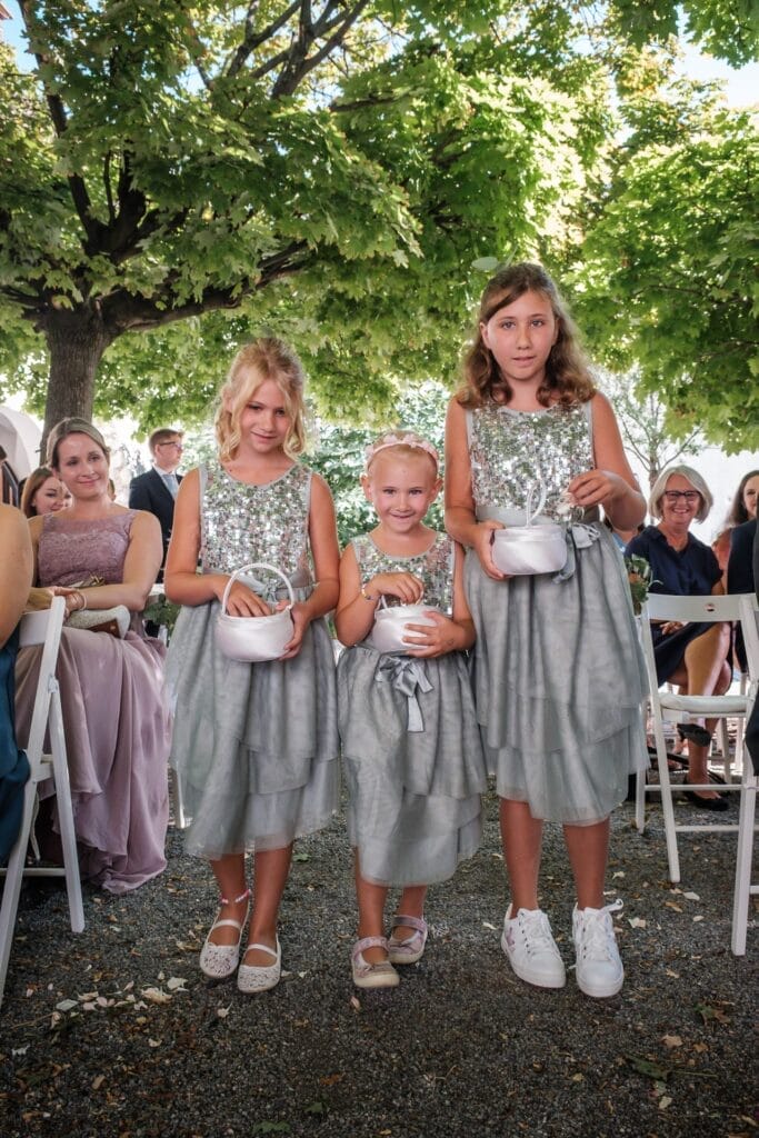 Hochzeitskrone Drei junge Mädchen in passenden silbernen Kleidern stehen mit Körben in der Hand bei einer Veranstaltung im Freien und fangen bezaubernde Momente mit den hinter ihnen sitzenden Gästen ein, ein perfektes Beispiel für natürliche Hochzeitsfotos.