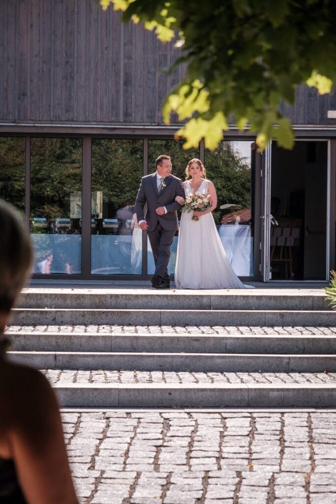 Hochzeitskrone Eine Braut in einem weißen Kleid wird von einem Mann im Anzug eine Steintreppe hinunter vor ein Gebäude mit großen Glastüren begleitet. Im Vordergrund ist eine Person zu sehen, die teilweise sichtbar bezaubernde Momente der Hochzeitsfotografie in Niederösterreich festhält.