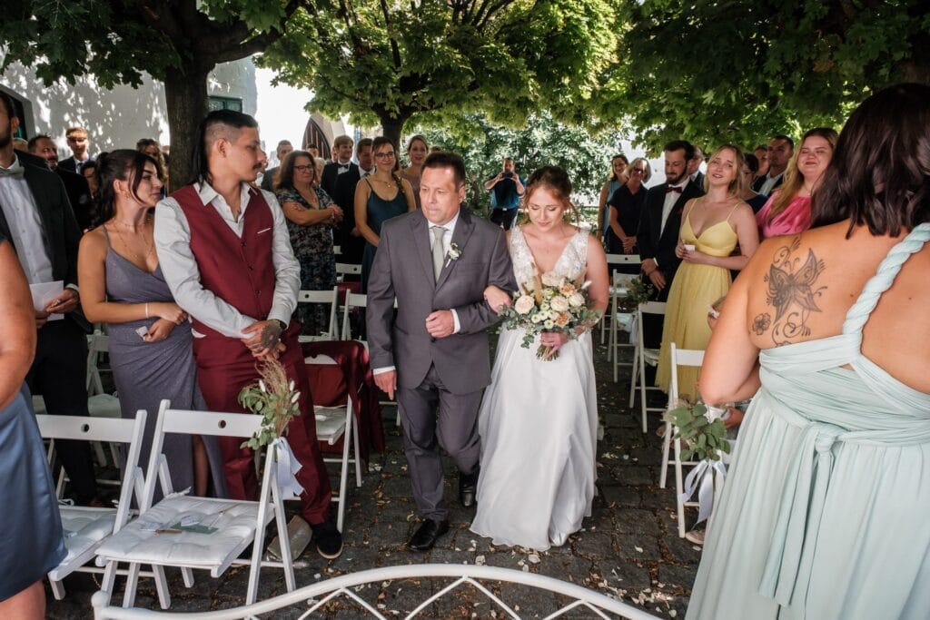 Hochzeitskrone Eine Braut schreitet mit einem Mann im Anzug den Gang entlang, umgeben von Gästen, die draußen unter Bäumen sitzen und bezaubernde Momente mit natürlichen Hochzeitsfotos festhalten.