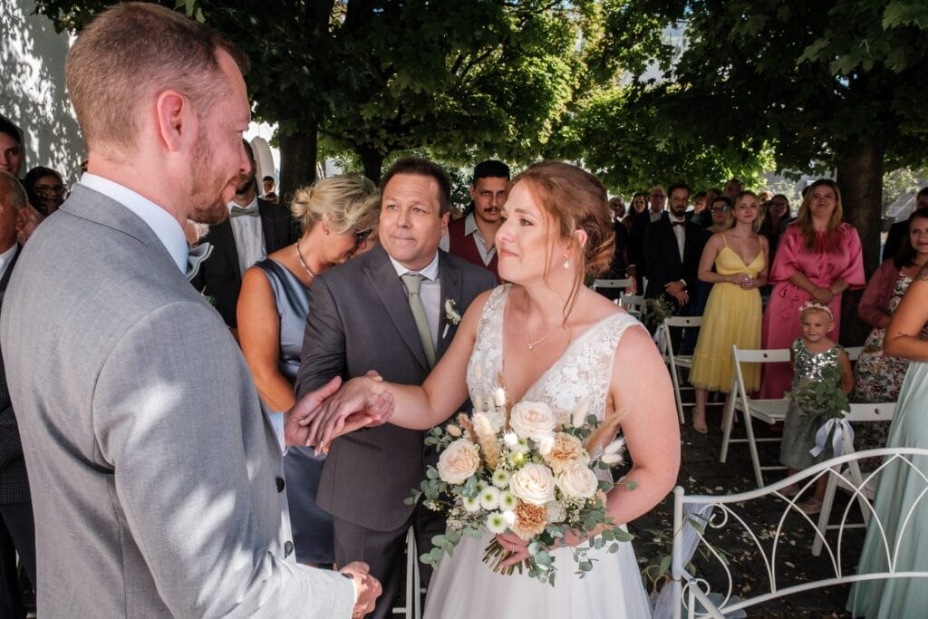 Hochzeitskrone Während einer Hochzeitszeremonie im Freien hält ein Paar Händchen, während die Braut einen Blumenstrauß in der Hand hält, umgeben von Gästen. Diese bezaubernden Momente eignen sich perfekt für natürliche Hochzeitsfotos und fangen die Essenz der Hochzeitsfotografie in Niederösterreich ein.