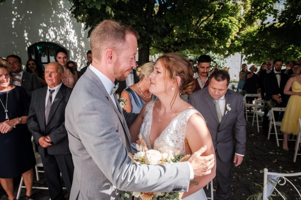 Hochzeitskrone Während der Hochzeitszeremonie umarmen sich Braut und Bräutigam unter freiem Himmel und halten bezaubernde Momente fest. Die Gäste schauen voller Ehrfurcht zu, wie diese natürlichen Hochzeitsfotos in Niederösterreich ihre Liebe verewigen.