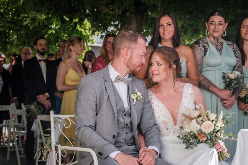 Hochzeitskrone Braut und Bräutigam sitzen eng beieinander unter einem Baum, umgeben von Brautjungfern und Gästen. Die Braut hält einen Blumenstrauß in der Hand und lächelt den Bräutigam in seinem hellgrauen Anzug an. Sie hält bezaubernde Momente ihres besonderen Tages in natürlichen Hochzeitsfotos fest.