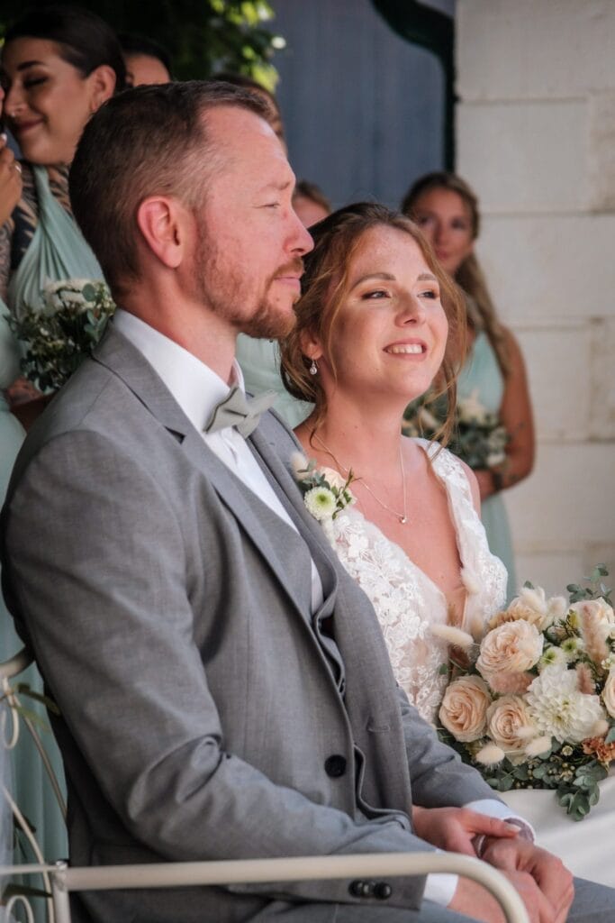 Hochzeitskrone Das in Hochzeitskleidung gekleidete Paar sitzt zusammen, während die Braut einen Blumenstrauß in der Hand hält. Brautjungfern in Pastellkleidern tragen zum Charme der Szene bei. Dieser Moment fängt bezaubernde Momente natürlicher Hochzeitsfotos ein und verkörpert Hochzeitsfotografie in Niederösterreich.