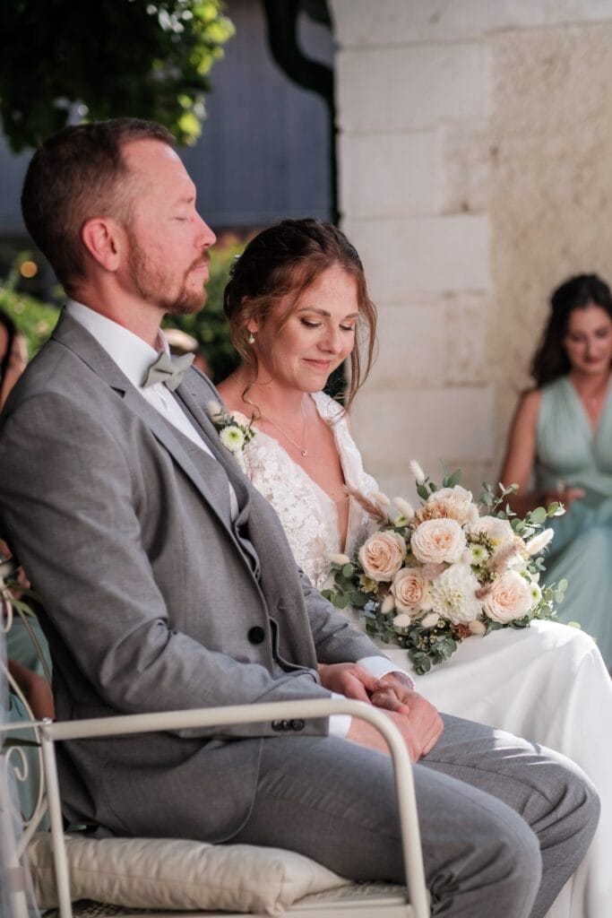 Hochzeitskrone Braut und Bräutigam sitzen zusammen, die Augen geschlossen, vielleicht während einer Hochzeitszeremonie, während die Braut einen Strauß blasser Rosen hält. Dieser Moment fängt die Essenz natürlicher Hochzeitsfotos ein und bewahrt bezaubernde Momente in zeitloser Schönheit.