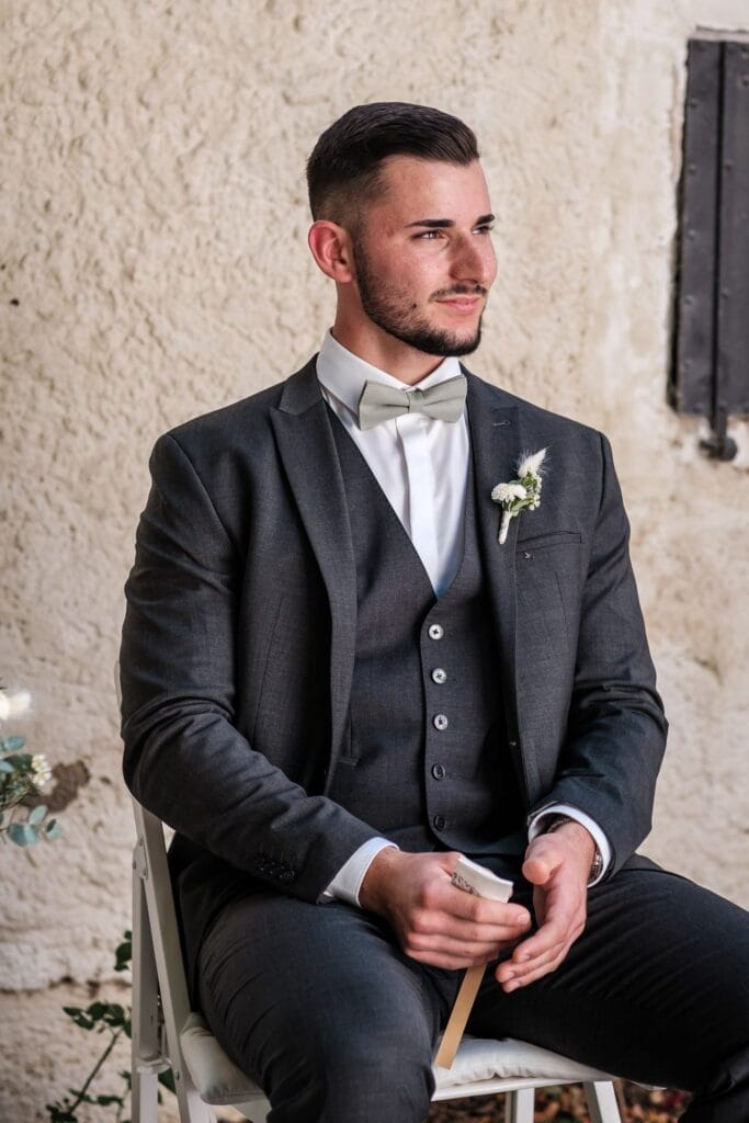 Hochzeitskrone Ein Mann in einem dunklen Anzug mit Weste und Fliege sitzt auf einem Stuhl und hält ein Blatt Papier in der Hand. Er trägt eine Ansteckblume am Revers und steht vor einer strukturierten Wand. Er fängt einen der bezaubernden Momente ein, die typisch für die Hochzeitsfotografie in Niederösterreich sind.