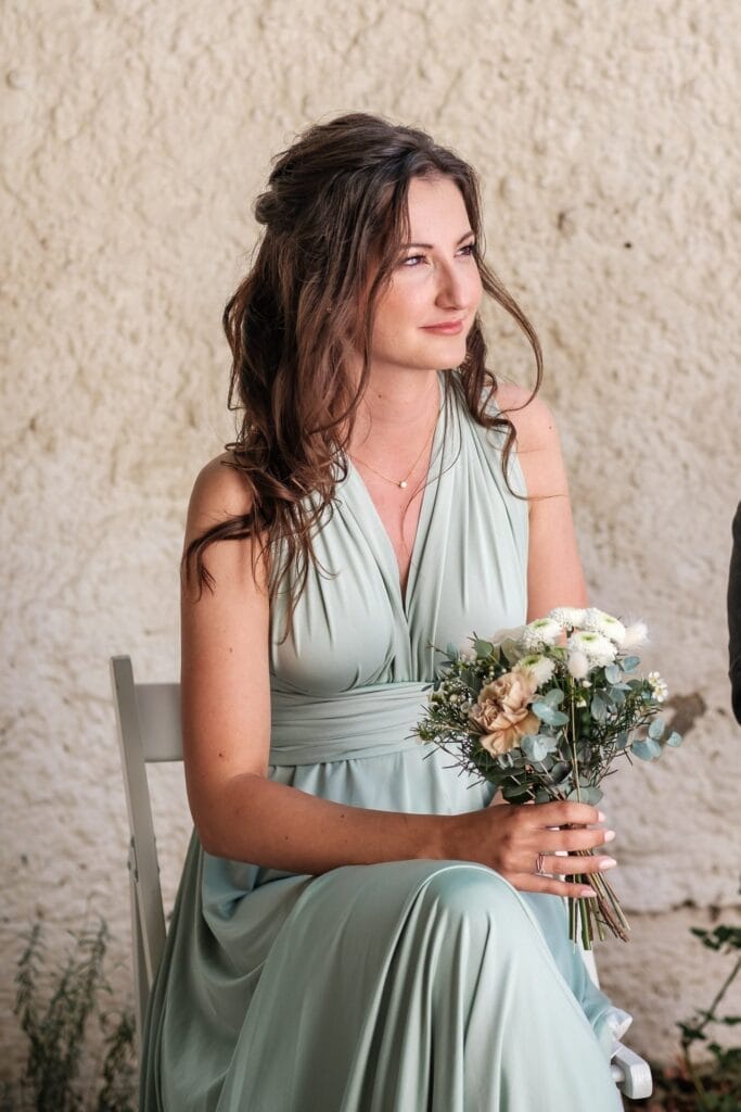 Hochzeitskrone Eine Frau in einem hellgrünen Kleid sitzt anmutig auf einem Stuhl, hält einen Blumenstrauß vor einer strukturierten Wand und fängt bezaubernde Momente ein.