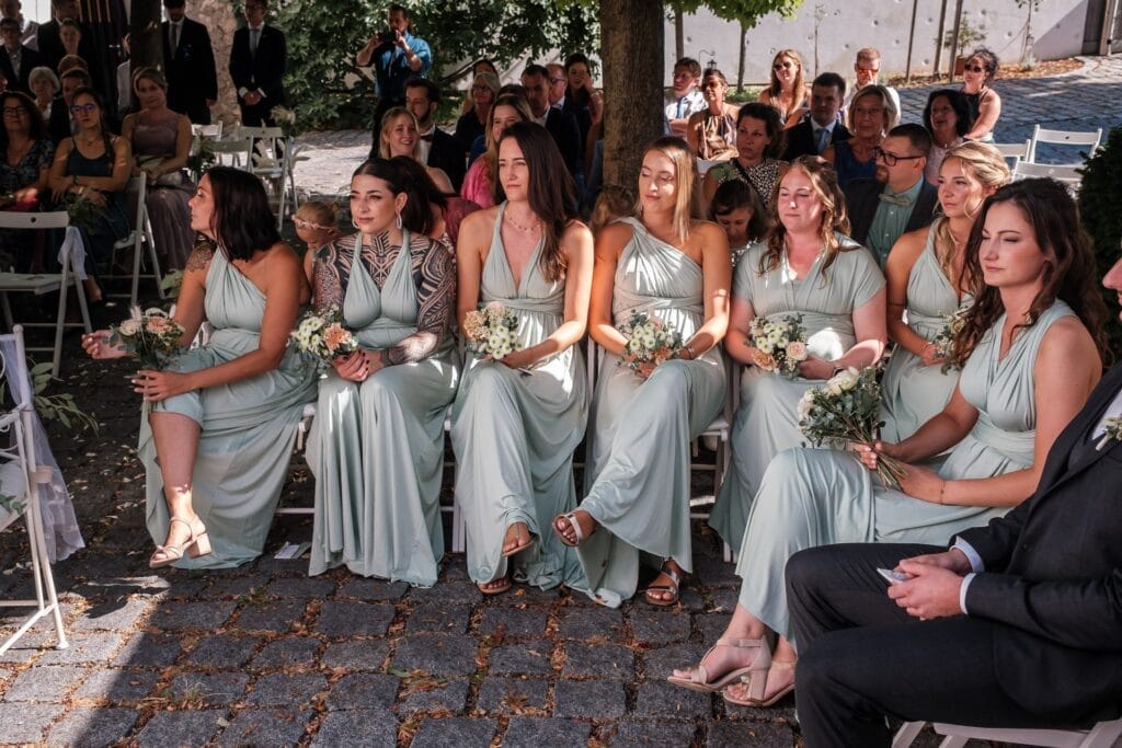 Hochzeitskrone Eine Gruppe Brautjungfern in passenden grünen Kleidern sitzt draußen auf weißen Stühlen und hält Blumensträuße in den Händen. Diese natürlichen Hochzeitsfotos fangen bezaubernde Momente ein, mit nachdenklich im Hintergrund sitzenden Gästen.