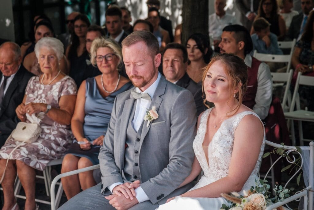 Hochzeitskrone Bei einer Hochzeitszeremonie im Freien in Niederösterreich sitzen Braut und Bräutigam zusammen, umgeben von sitzenden Gästen. Die Szene fängt bezaubernde Momente ein, die natürliche Hochzeitsfotos ausmachen und die Essenz ihres besonderen Tages bewahren.