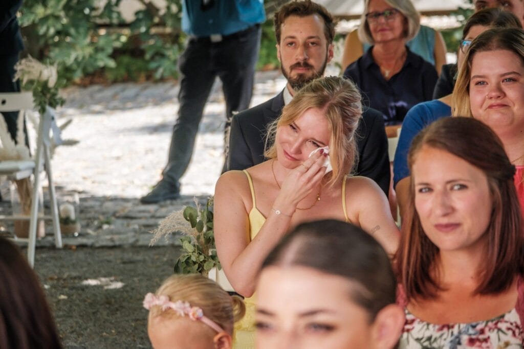 Hochzeitskrone Eine Frau in einem gelben Kleid wischt sich Tränen aus den Augen und fängt einen dieser bezaubernden Momente inmitten der Menschenmenge bei einer Veranstaltung im Freien ein.