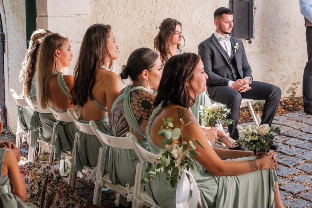 Hochzeitskrone Eine Gruppe Brautjungfern in grünen Kleidern und ein Mann im Anzug sitzen bei einer Hochzeitszeremonie auf weißen Stühlen und halten bezaubernde Momente in natürlichen Hochzeitsfotos fest.