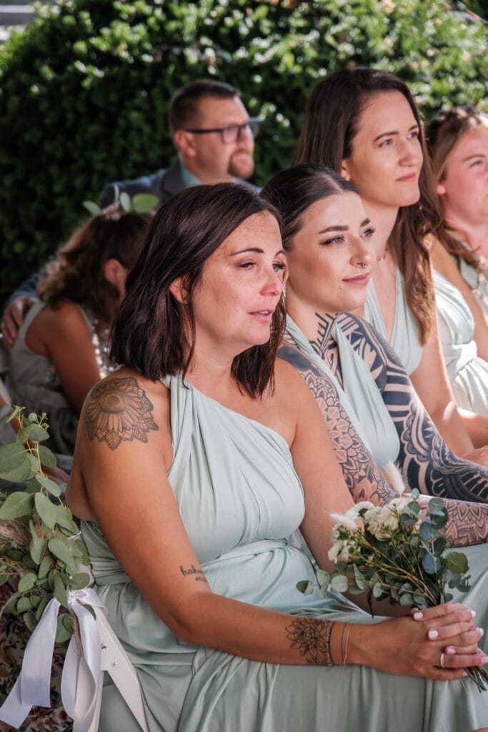 Hochzeitskrone Frauen in hellgrünen Kleidern, geschmückt mit Tätowierungen, halten Blumen in der Hand und beobachten aufmerksam ein Ereignis. Diese natürlichen Hochzeitsfotos fangen bezaubernde Momente der Anmut und Vorfreude ein und verkörpern die Essenz der Hochzeitsfotografie in Niederösterreich.