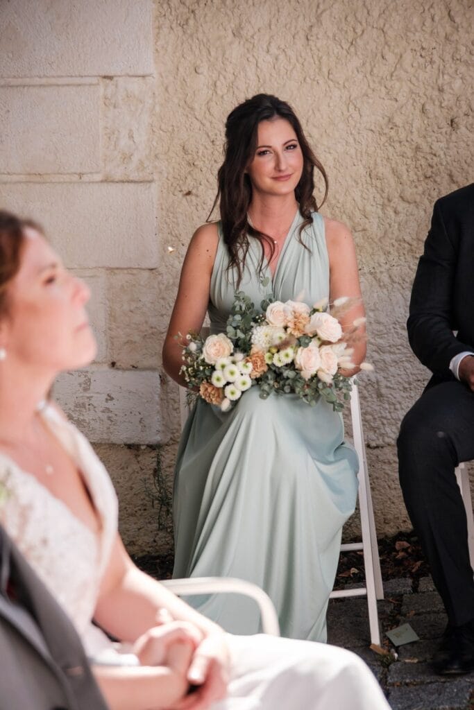 Hochzeitskrone Eine Frau in einem grünen Kleid, die einen Blumenstrauß hält, sitzt im Freien neben einer Person in einem weißen Outfit und fängt bezaubernde Momente ein. Perfekt für diejenigen, die natürliche Hochzeitsfotos suchen, die authentische Freude und Verbundenheit zeigen.