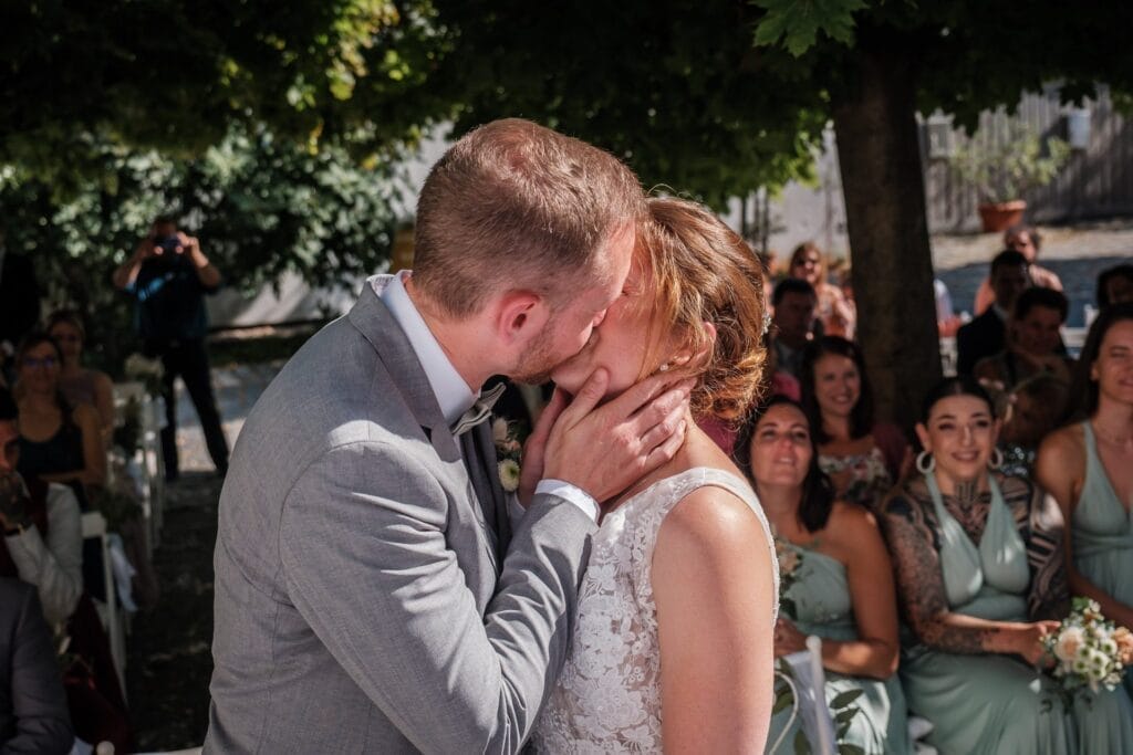 Hochzeitskrone Ein Paar küsst sich bei einer Hochzeitszeremonie im Freien, umgeben von sitzenden Gästen. Der Bräutigam trägt einen grauen Anzug und die Braut ein weißes Kleid – bezaubernde Momente festgehalten in natürlichen Hochzeitsfotos.