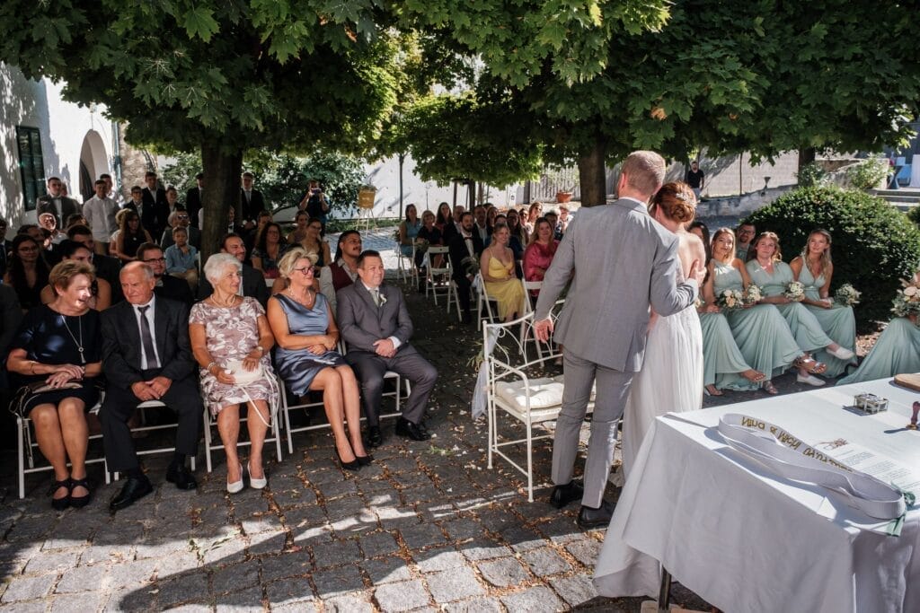 Hochzeitskrone Ein Paar steht bei einer Hochzeitszeremonie im Freien in Niederösterreich und blickt unter sonnenbeschienenen Bäumen auf seine Gäste. Das Sonnenlicht fällt sanft durch die Blätter und diese bezaubernden Momente werden festgehalten. Im Vordergrund steht ein weißer Tisch mit Gegenständen, der den Charme natürlicher Hochzeitsfotos verkörpert.