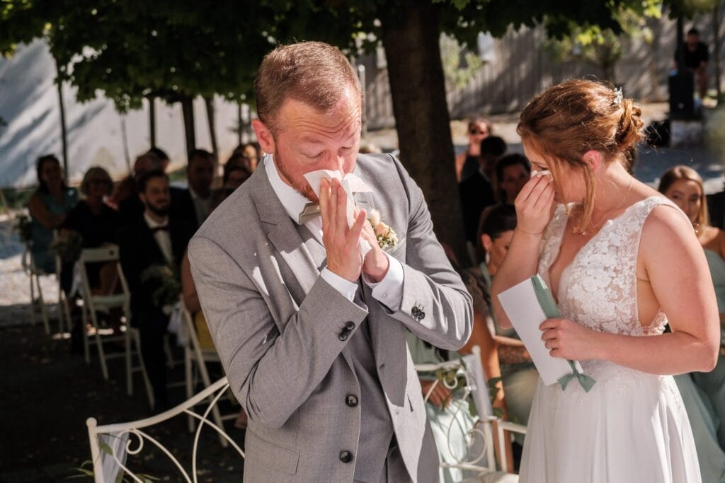 Hochzeitskrone Braut und Bräutigam, beide emotional, sind in bezaubernden Momenten während einer Hochzeitszeremonie im Freien festgehalten. Der Bräutigam trägt einen grauen Anzug und die Braut ein weißes Kleid, beide wischen sich die Augen.