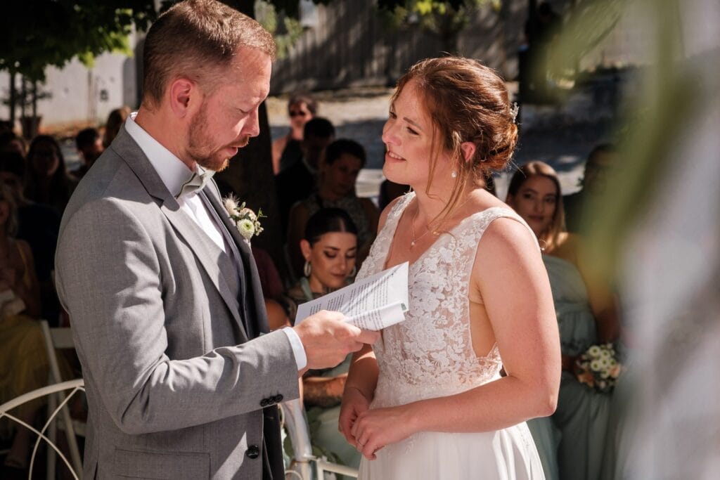 Hochzeitskrone Ein Paar steht während seiner Hochzeitszeremonie, festgehalten in bezaubernden Momenten. Der Bräutigam, im grauen Anzug, hält Papiere, während die Braut in Weiß ihn anschaut. Im Hintergrund sitzen die Gäste, alle wunderschön eingerahmt von natürlichen Hochzeitsfotos.