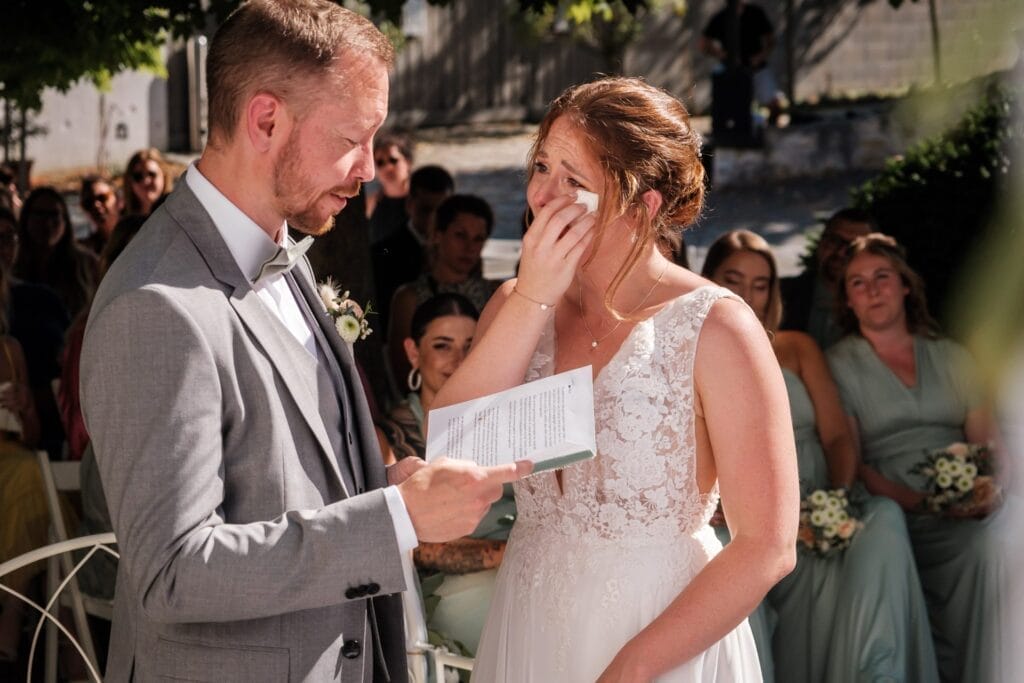 Hochzeitskrone Ein Mann im grauen Anzug liest aus einer Zeitung und fängt bezaubernde Momente ein, während eine Frau im weißen Kleid sich die Augen wischt. Diese Szene natürlicher Hochzeitsfotos spielt sich im Freien ab, mit sitzenden Menschen im Hintergrund, und ist ein Sinnbild für Hochzeitsfotografie in Niederösterreich.