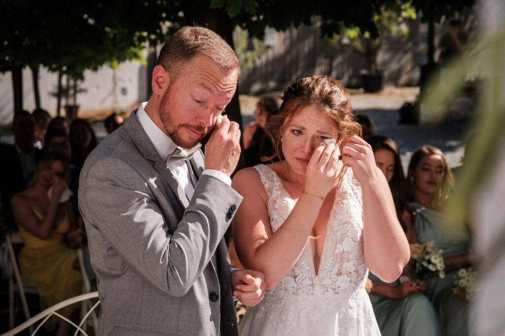 Hochzeitskrone Braut und Bräutigam in Abendgarderobe stehen Seite an Seite und wischen sich Tränen aus den Augen in einer Darstellung bezaubernder Momente, während im Hintergrund die Hochzeitsgäste natürliche Hochzeitsfotos schießen.