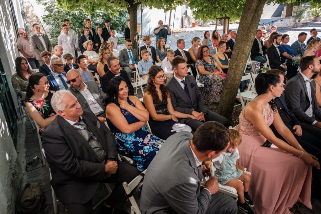 Hochzeitskrone Inmitten der ruhigen Baumkulisse versammelte sich eine Gruppe in eleganten Anzügen und Kleidern zu einem formellen Anlass. Ein Kind sitzt auf dem Schoß eines Mannes und fängt bezaubernde Momente ein, die sich perfekt für natürliche Hochzeitsfotos eignen.