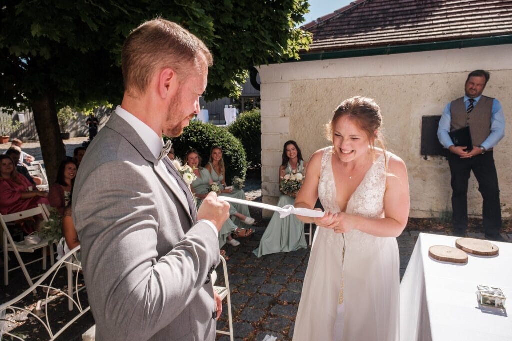 Hochzeitskrone Inmitten üppigen Grüns nehmen Braut und Bräutigam an einer fröhlichen Handfasting-Zeremonie teil. Die ruhige Umgebung bietet die perfekte Kulisse für bezaubernde Momente, die in natürlichen Hochzeitsfotos festgehalten werden, während die Gäste rundherum sitzen und ihre Verbindung feiern.