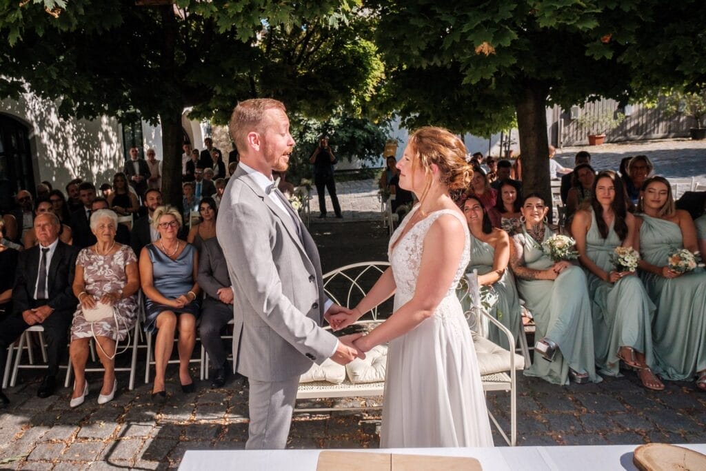 Hochzeitskrone Inmitten der bezaubernden Kulisse einer Zeremonie im Freien stehen Braut und Bräutigam Händchen haltend, umgeben von sitzenden Gästen. Diese bezaubernden Momente werden wunderschön durch natürliche Hochzeitsfotos festgehalten, die die Eleganz der Hochzeitsfotografie in Niederösterreich widerspiegeln.