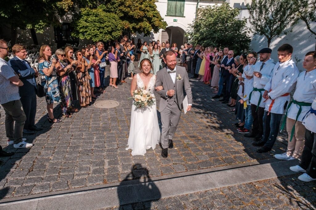 Hochzeitskrone Braut und Bräutigam gehen Arm in Arm einen Kopfsteinpflasterweg entlang und halten bezaubernde Momente fest, während die Menge applaudiert und Fotos macht.