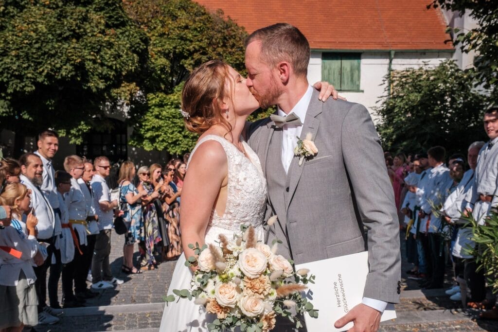 Hochzeitskrone Inmitten einer Menschenmenge küssen sich Braut und Bräutigam im Freien. Sie umklammert ihren Blumenstrauß, während er ein Dokument hält. So werden bezaubernde Momente vor dem Hintergrund üppigen Grüns festgehalten. Es ist ein authentisches Beispiel für Hochzeitsfotografie in Niederösterreich.