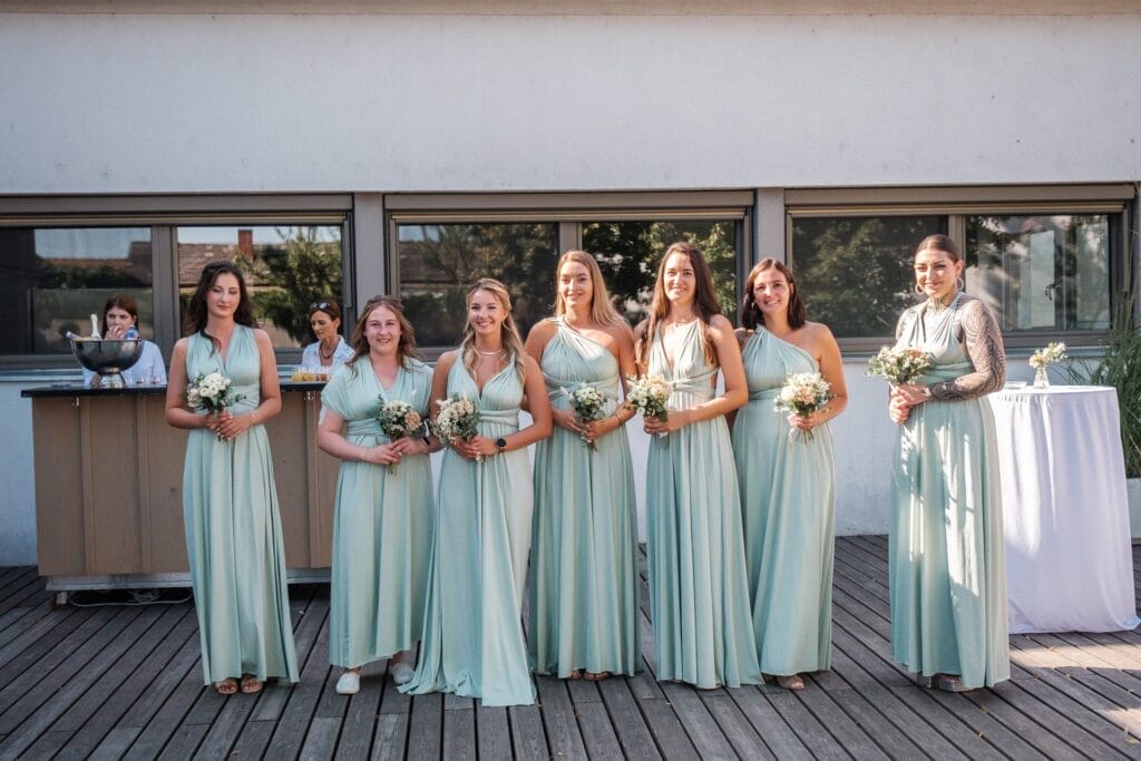 Hochzeitskrone Sieben Brautjungfern in hellgrünen Kleidern stehen in einer Reihe und halten Blumensträuße auf einem Holzdeck. Im Hintergrund sind eine Bar und Menschen zu sehen. Das Einfangen dieser bezaubernden Momente ist ein Beispiel exquisiter Hochzeitsfotografie in Niederösterreich.