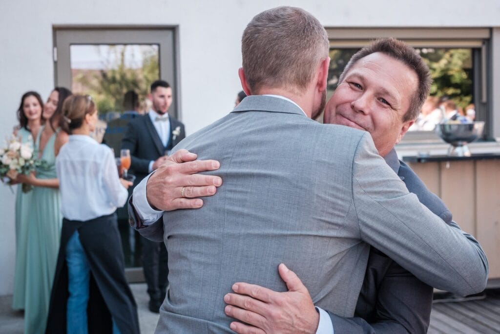 Hochzeitskrone Inmitten einer lebhaften Versammlung umarmen sich zwei Männer in Anzügen und fangen vor dem Hintergrund freudiger Zuschauer einen dieser bezaubernden Momente ein.