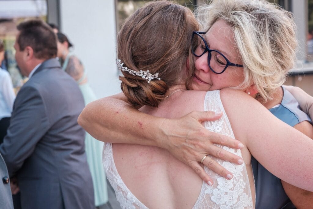 Hochzeitskrone Zwei Frauen umarmen sich herzlich bei einer Hochzeit, eine im Spitzenkleid und mit Haarteil, die andere mit Brille und kurzen Haaren. Um sie herum entfalten sich Bezaubernde Momente der Hochzeitsfotografie in Niederösterreich, während Gäste in festlicher Kleidung den Hintergrund füllen.