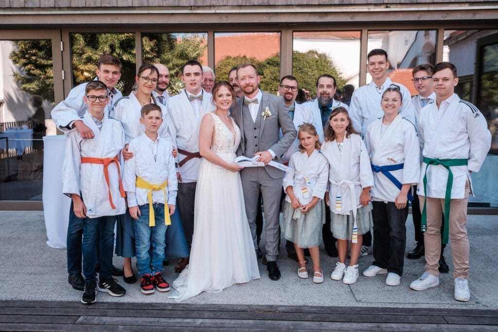 Hochzeitskrone Braut und Bräutigam stehen zusammen, festgehalten in bezaubernde Momente von Hochzeitsfotografie in Niederösterreich. Sie posieren im Freien, umgeben von Menschen in Kampfsportuniformen mit Gürteln in verschiedenen Farben, vor einem großen Gebäude mit Glasfenstern.