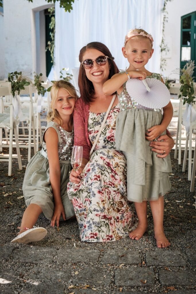 Hochzeitskrone Eine Frau kniet zwischen zwei jungen Mädchen in einer ruhigen Umgebung im Freien, umgeben von weißen Stühlen. In diesem Moment natürlicher Hochzeitsfotos hält sie ein Glas, während die Mädchen in passenden silbernen und grünen Kleidern bezaubernde Momente festhalten.