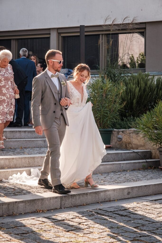 Hochzeitskrone Eine Braut und ein Bräutigam gehen lächelnd die Außentreppe hinunter und erleben bezaubernde Momente. Die Braut hält ihr Kleid in der Hand und trägt so zur natürlichen Hochzeitsfotos-Atmosphäre bei. Im Hintergrund verweilen die Gäste und fangen die Essenz der Hochzeitsfotografie in Niederösterreich ein.