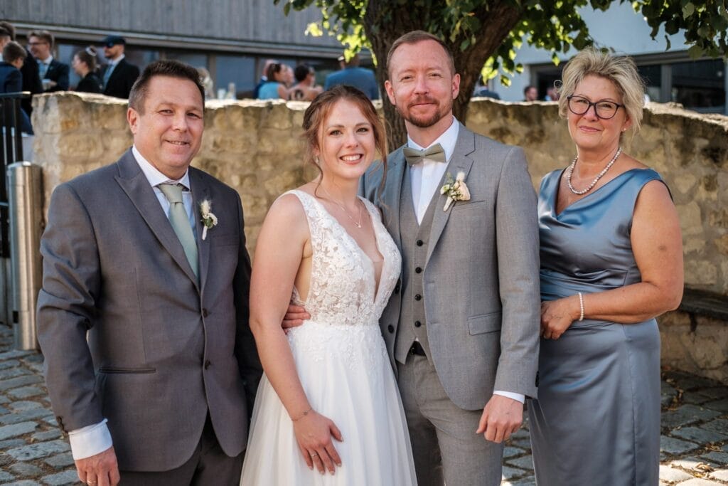 Hochzeitskrone Vier Personen posieren gemeinsam im Freien in formeller Kleidung, umgeben von einem Baum und einer Steinmauer, und fangen bezaubernde Momente ein. Dies ist ein perfektes Beispiel für natürliche Hochzeitsfotos, die den Charme der Hochzeitsfotografie in Niederösterreich wirklich widerspiegeln.