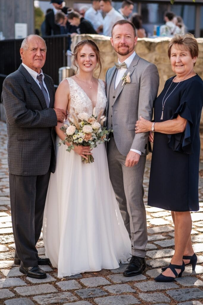 Hochzeitskrone Braut und Bräutigam posieren im Freien mit einem älteren Paar und halten bezaubernde Momente fest. Die Braut hält einen Blumenstrauß, der Bräutigam trägt einen hellgrauen Anzug, während der ältere Mann einen Anzug und die Frau ein marineblaues Kleid trägt – perfekte natürliche Hochzeitsfotos in Niederösterreich.