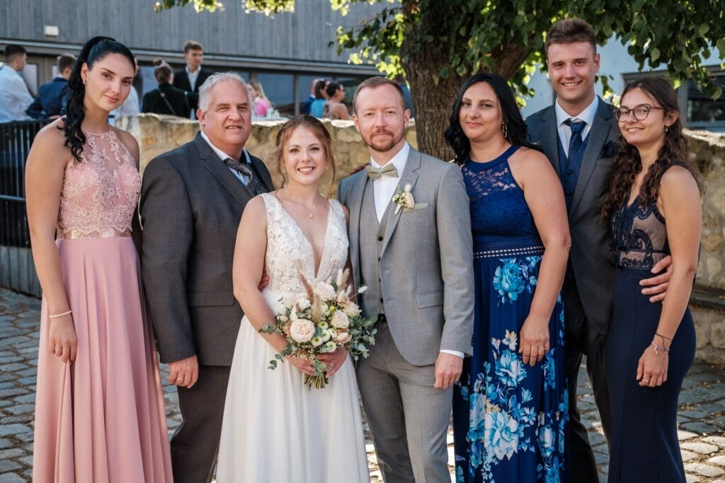 Hochzeitskrone Ein Hochzeitsgruppenfoto mit sieben Personen fängt bezaubernde Momente ein. Braut und Bräutigam stehen in der Mitte, umgeben von drei Frauen und zwei Männern, und fangen so natürliche Hochzeitsfotos ein. Sie stehen draußen an einem sonnigen Tag neben einem Baum und sind ein Beispiel für Hochzeitsfotografie in Niederösterreich.