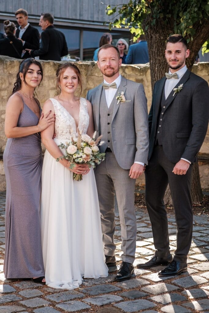 Hochzeitskrone Vier Personen in formeller Kleidung stehen zusammen im Freien: eine Braut in einem weißen Kleid, ein Bräutigam in einem grauen Anzug und zwei Gäste. Sie stehen auf einem Steinweg mit einem Baum in der Nähe und fangen bezaubernde Momente der Hochzeitsfotografie in Niederösterreich ein.