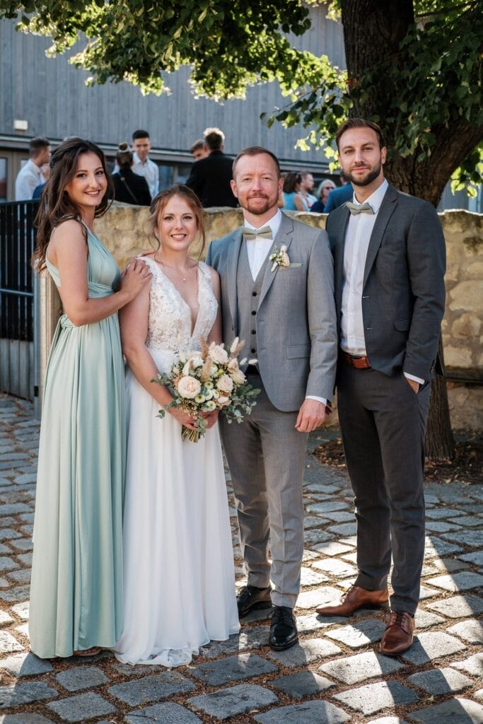 Hochzeitskrone Braut und Bräutigam stehen zusammen mit zwei gut gekleideten Gästen an einem sonnigen Tag im Freien vor einem Baum und einem Gebäude und halten bezaubernde Momente in der Hochzeitsfotografie in Niederösterreich fest.