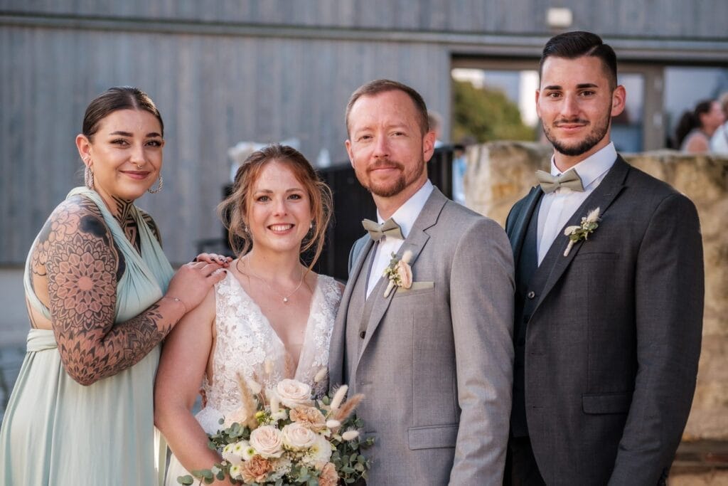 Hochzeitskrone Vier Personen in formeller Kleidung posieren gemeinsam bei einer Veranstaltung im Freien und halten bezaubernde Momente fest. Zwei Frauen tragen Kleider und zwei Männer Anzüge. Die Frau in Weiß hält einen Blumenstrauß in der Hand, ein Sinnbild für natürliche Hochzeitsfotos.