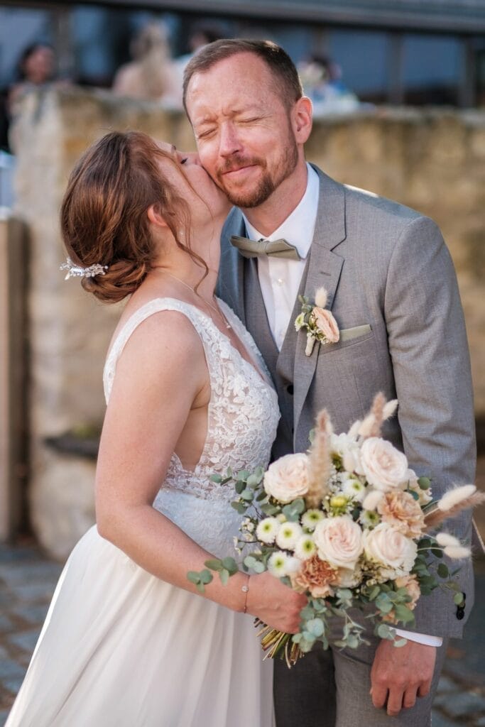 Hochzeitskrone In bezaubernden Momenten der Hochzeitsfotografie in Niederösterreich küsst die Braut im weißen Kleid den Bräutigam im grauen Anzug auf die Wange. Er hält einen Strauß aus weißen und pfirsichfarbenen Blumen und strahlt vor Freude.