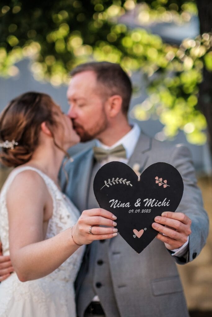 Hochzeitskrone Ein Paar küsst sich und hält ein schwarzes herzförmiges Schild mit Blumen und dem Text „Nina & Michi, 09.09.2025“ in der Hand und hält bezaubernde Momente fest. Sie sind draußen mit Bäumen im Hintergrund, perfekt für natürliche Hochzeitsfotos in Niederösterreich.