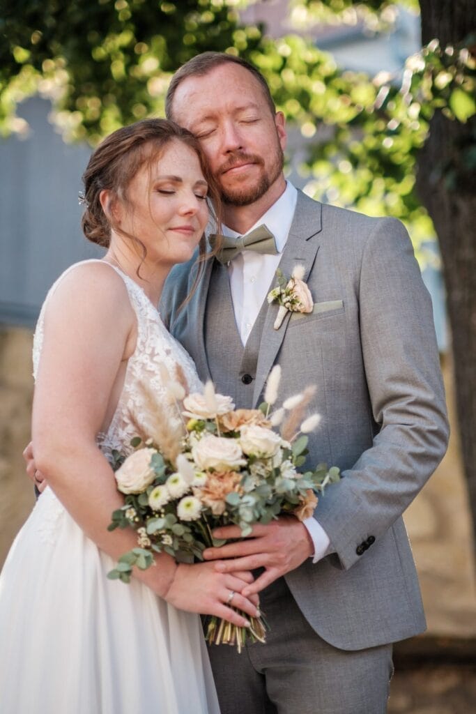 Hochzeitskrone Ein Paar teilt an einem sonnigen Tag einen bezaubernden Moment im Freien. Die Frau hält einen Blumenstrauß in der Hand, beide in formeller Kleidung gekleidet, und fängt natürliche Hochzeitsfotos ein, die die Eleganz der Hochzeitsfotografie in Niederösterreich widerspiegeln.