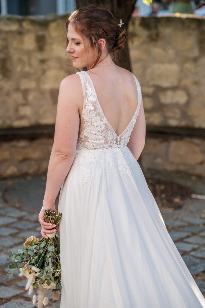 Hochzeitskrone Die Braut, die ein weißes Spitzenbrautkleid trägt und einen Blumenstrauß hält, steht anmutig auf einem Kopfsteinpflasterweg. Dieser Moment fängt die Essenz natürlicher Hochzeitsfotos ein und bewahrt bezaubernde Momente der Liebe und Freude.