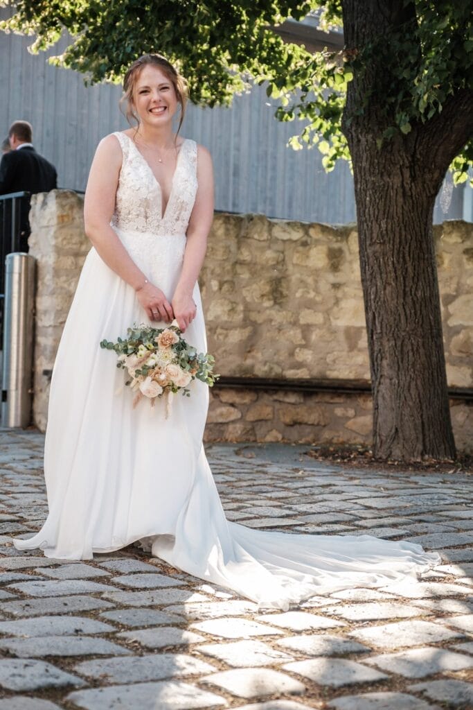 Hochzeitskrone Eine lächelnde Braut in einem weißen Kleid hält einen Blumenstrauß auf einem Kopfsteinpflasterweg neben einem Baum und einer Steinmauer und fängt bezaubernde Momente natürlicher Hochzeitsfotos ein.