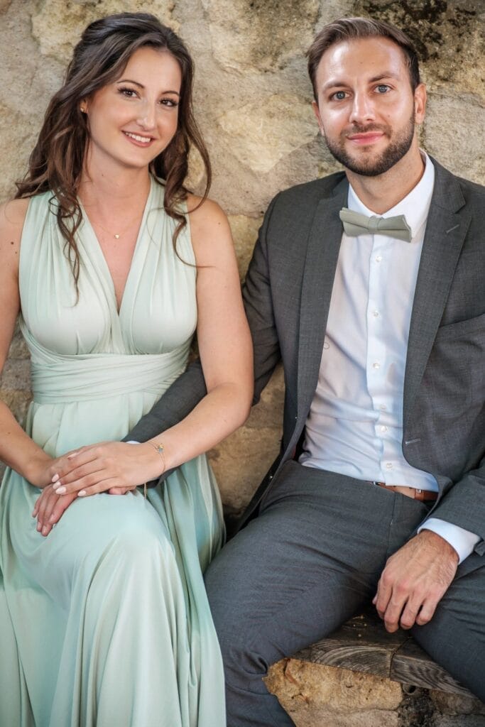 Hochzeitskrone Eine Frau in einem hellgrünen Kleid und ein Mann in einem grauen Anzug mit grüner Fliege sitzen lächelnd zusammen und fangen bezaubernde Momente ein.