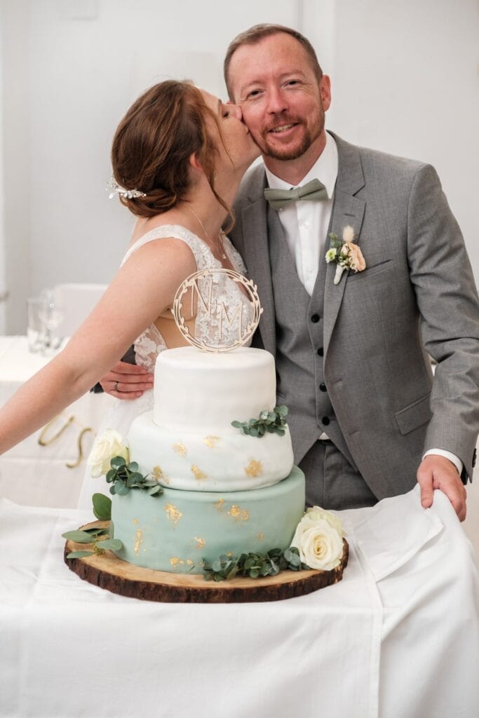 Hochzeitskrone Die Braut im weißen Kleid küsst den Bräutigam im grauen Anzug neben einer gestuften Hochzeitstorte, geschmückt mit grünen und goldenen Akzenten, und fängt bezaubernde Momente natürlicher Hochzeitsfotos für Ihre perfekte Hochzeitsfotografie in Niederösterreich ein.