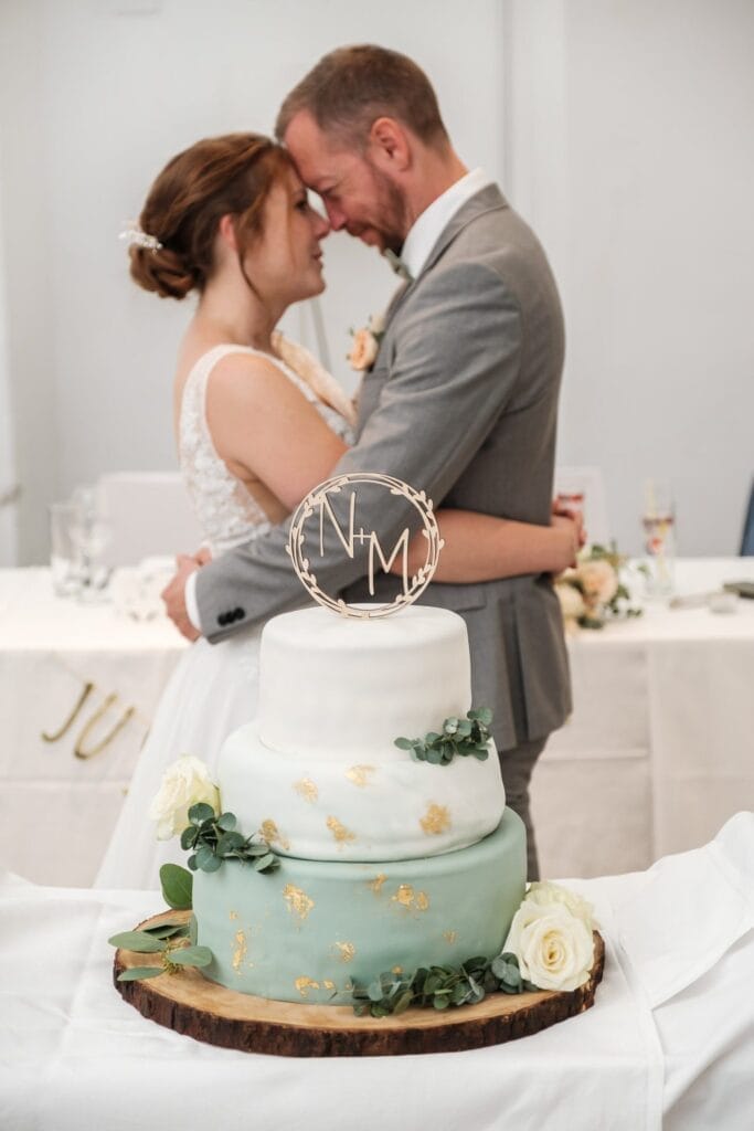 Hochzeitskrone Braut und Bräutigam teilen bezaubernde Momente, Stirn an Stirn umarmend hinter einer zweistöckigen Hochzeitstorte, die mit goldenen Akzenten und Grün geschmückt ist und wunderschön in natürlichen Hochzeitsfotos festgehalten wurde.