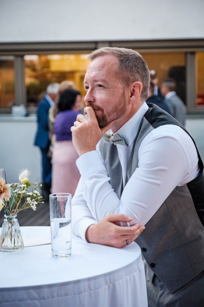 Hochzeitskrone Ein Mann in Weste und Krawatte sitzt nachdenklich an einem Tisch mit einem Glas Wasser und einer kleinen Vase mit Blumen und fängt bezaubernde Momente ein, die wie ein natürlicher Hochzeitsfoto wirken.