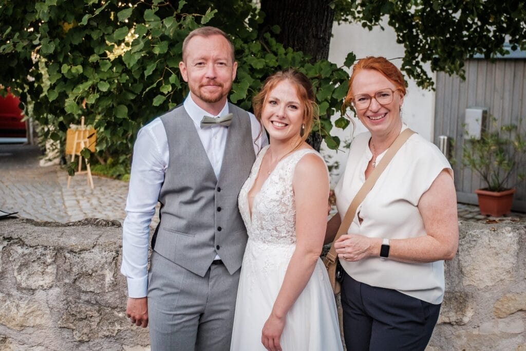 Hochzeitskrone Drei Personen posieren im Freien; ein Mann in einer Weste, eine Frau in einem Hochzeitskleid und eine Frau in einer Bluse mit Brille. Sie stehen vor einer Steinmauer mit Bäumen im Hintergrund und fangen bezaubernde Momente natürlicher Hochzeitsfotos ein.